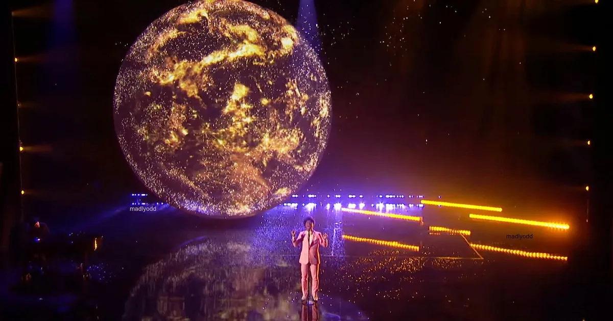 A person stands on a reflective stage with a large illuminated Earth backdrop and colorful stage lights.