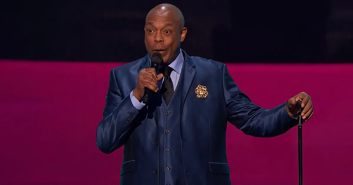 A man in a blue suit holds a microphone, gesturing with his other hand, against a pink background.