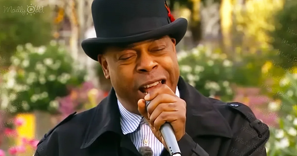 A man in a black coat and top hat sings into a microphone, surrounded by blurred flowers and greenery.