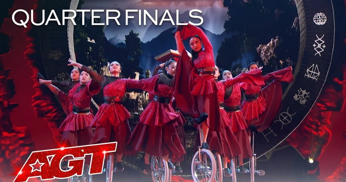 A performer in red balances on a unicycle, surrounded by dancers in matching red costumes against a scenic backdrop.