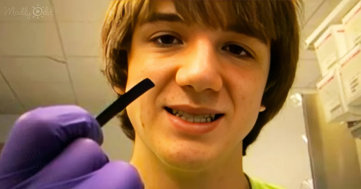 A person wearing purple gloves holds a pen near their mouth, with a tiled wall and shelves in the background.