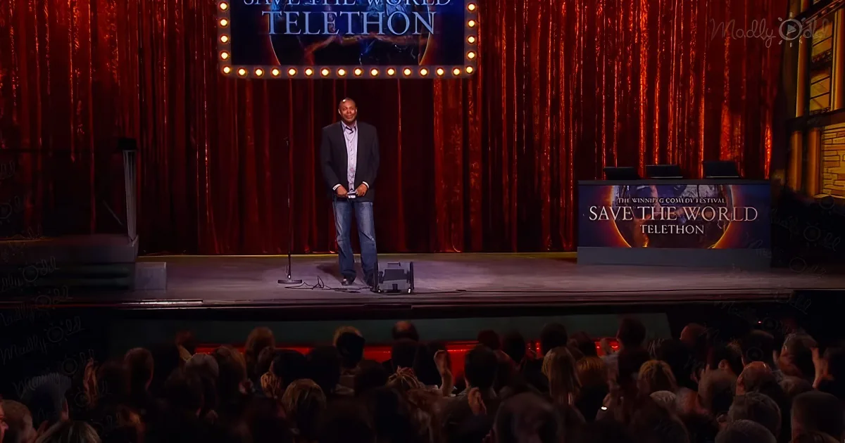 A man stands on stage at a "Save the World Telethon" event, addressing an audience.