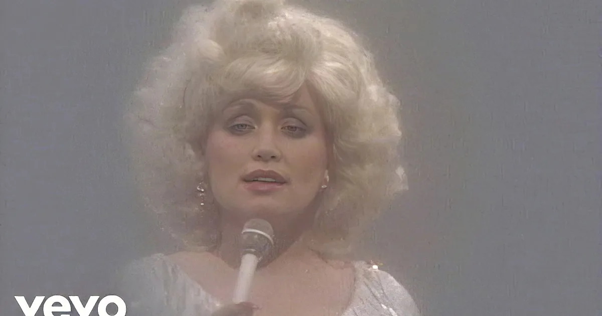 A person with blonde, curly hair and a white outfit sings into a microphone, surrounded by a hazy, ethereal effect.