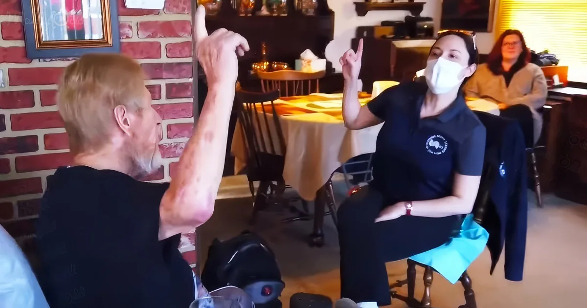 A man and a woman in a restaurant, both pointing upwards, with a brick wall and tables in the background.