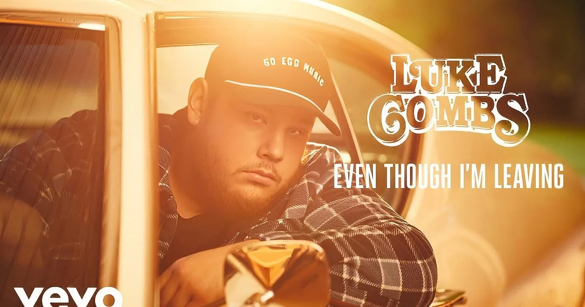 A man in a plaid shirt and cap sits in a car, looking out the window with a serious expression.