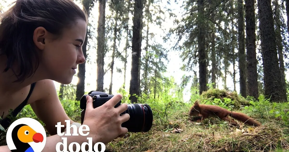 A woman smiles while holding a camera, surrounded by a forest, as a squirrel eats nearby.