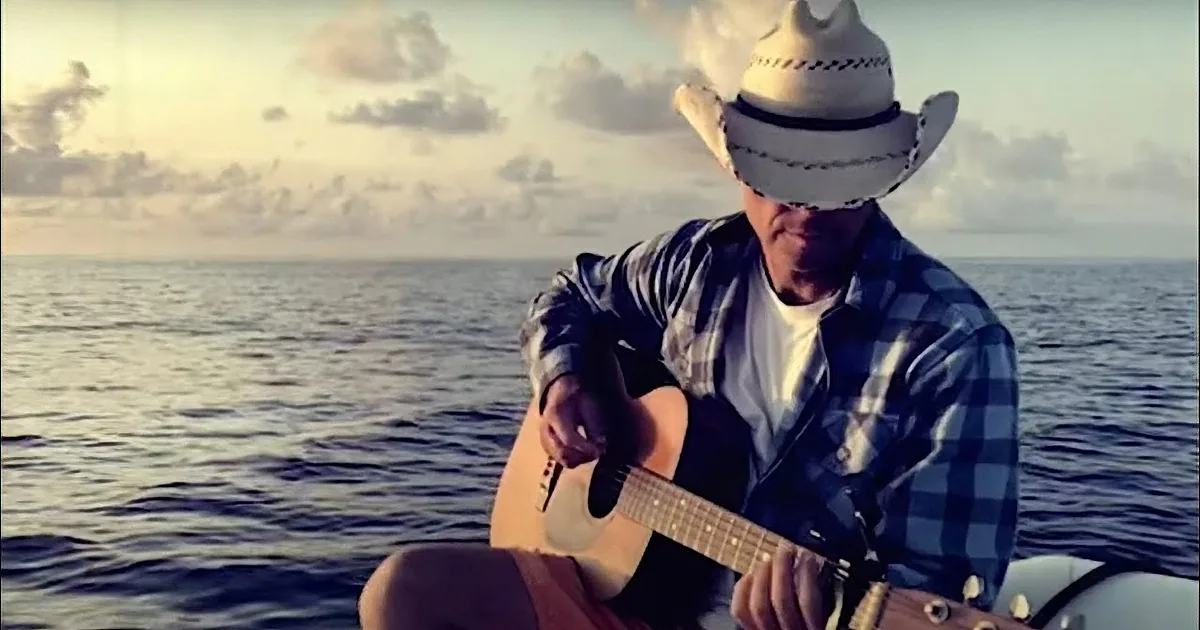 A man in a cowboy hat and plaid shirt plays guitar on a boat at sunset.