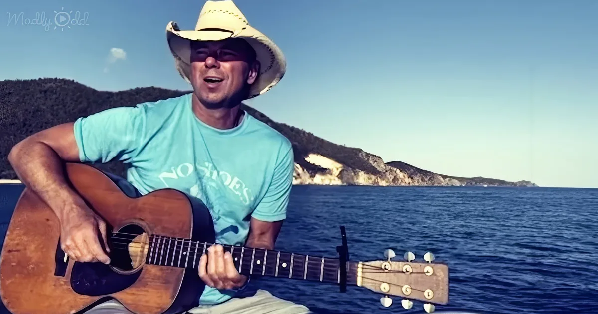 A man in a cowboy hat plays an acoustic guitar by the sea, with hills in the background.