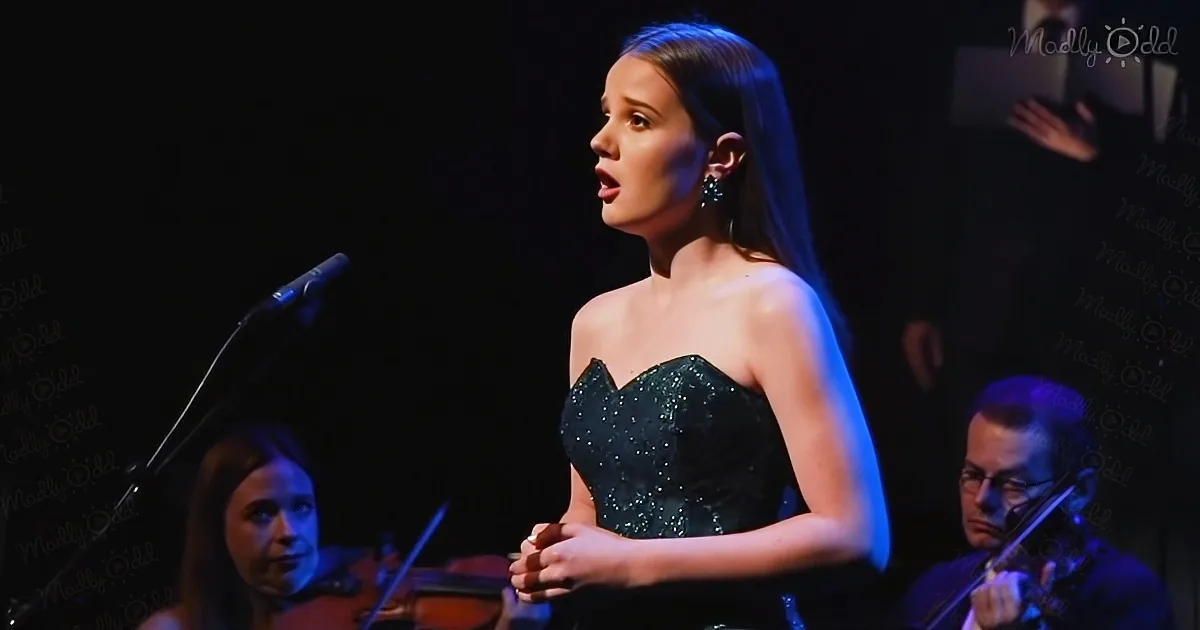 A woman in a black dress sings on stage with musicians playing violins behind her.