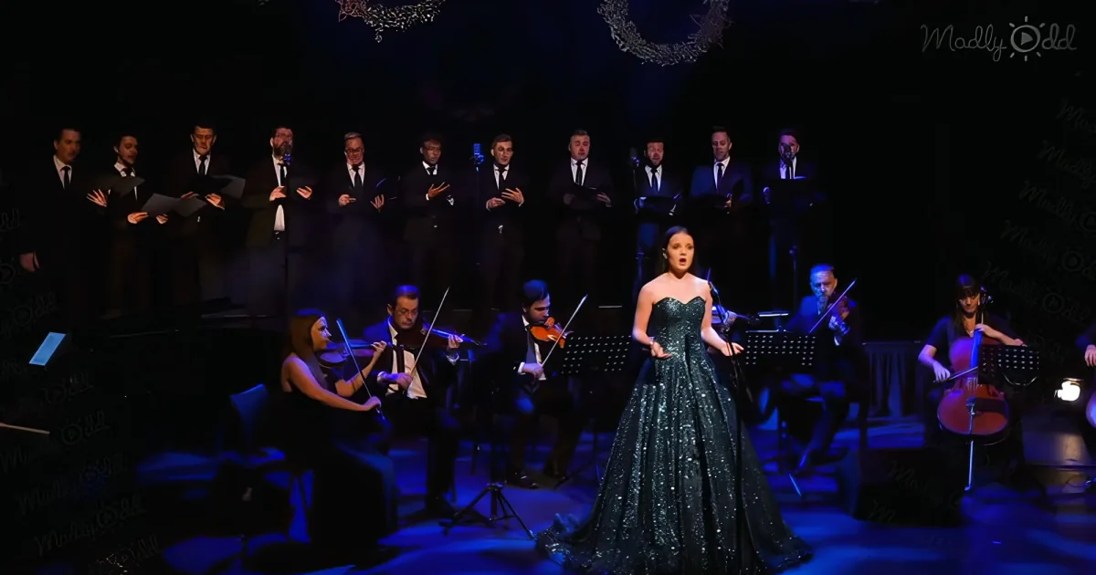 A woman in a sparkling dress sings on stage with a choir and orchestra behind her.