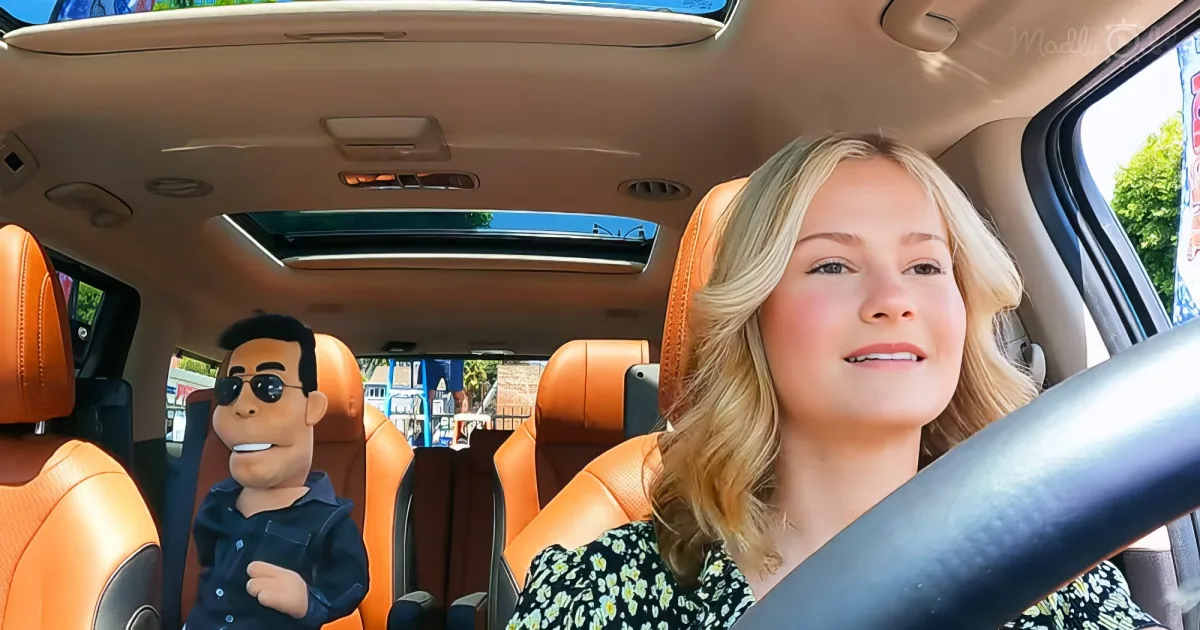 A woman drives a car with orange seats, smiling, while a cartoon character sits in the passenger seat.