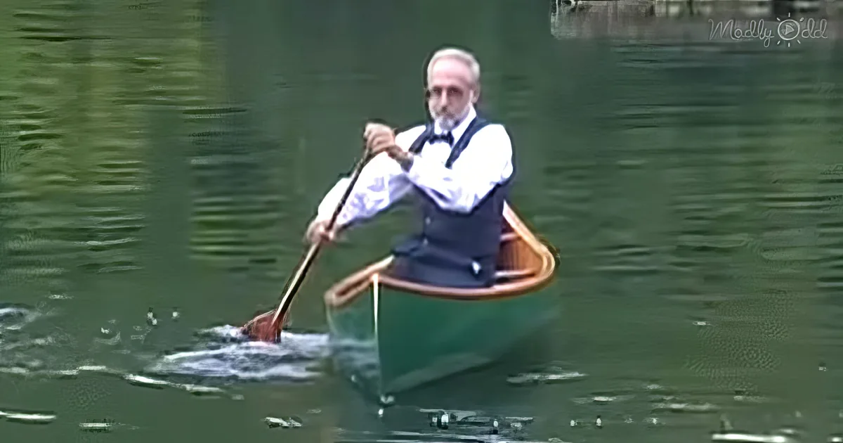 An older man in a white shirt and vest paddles a canoe on calm water.