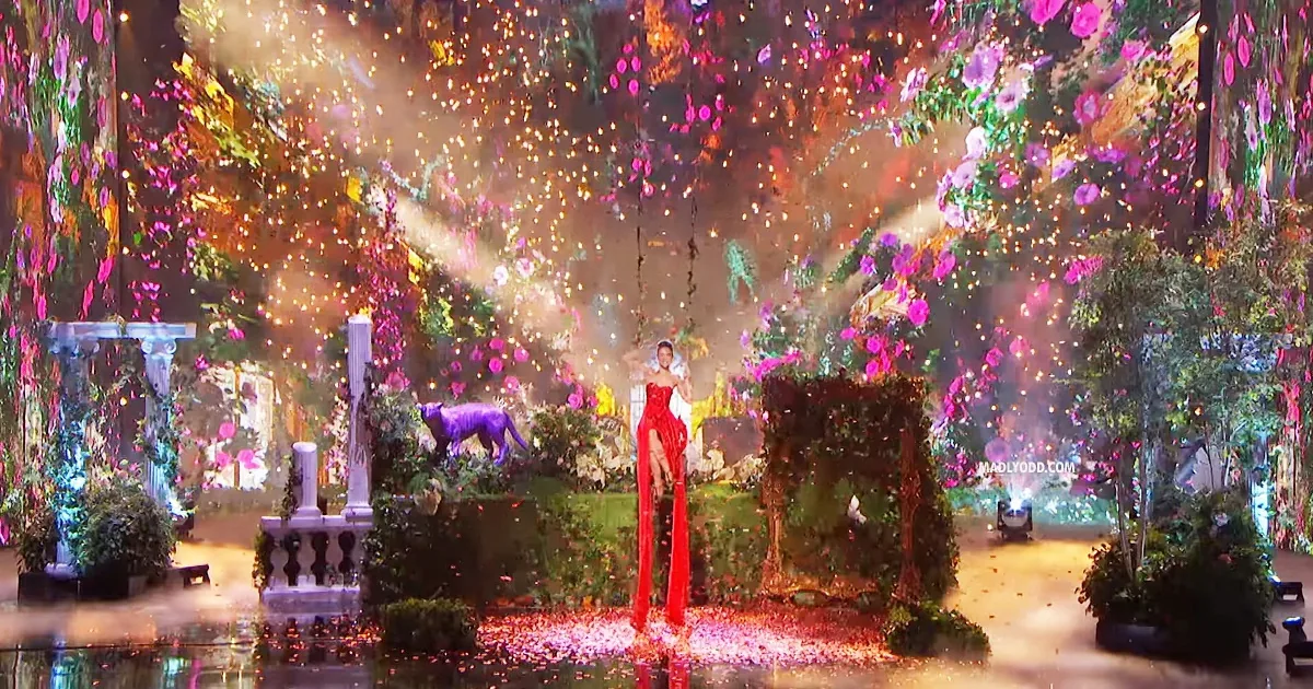 A woman in a red dress stands on stilts amidst a vibrant, flower-filled garden with falling petals and colorful lights.