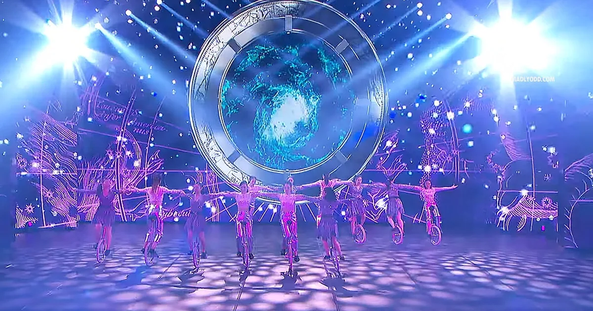 A group of performers on unicycles with arms outstretched, illuminated by vibrant stage lights and a large circular backdrop.