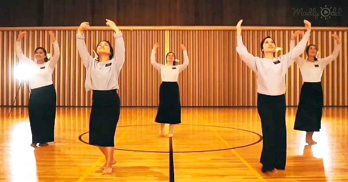 Four women in white tops and black skirts perform a synchronized dance on a wooden gym floor.