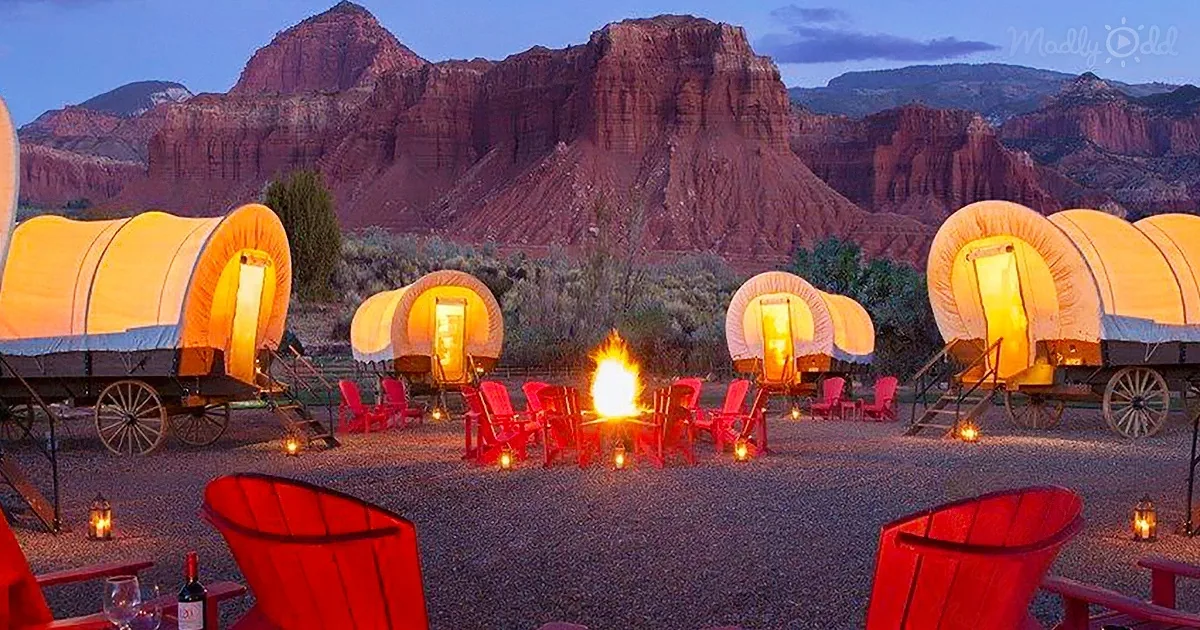 Amber-lit covered wagons surround a campfire with red chairs, set against a backdrop of rugged red rock mountains.