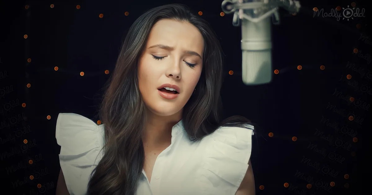 A woman with long dark hair sings into a microphone, wearing a white blouse, with a dark background and glowing lights.