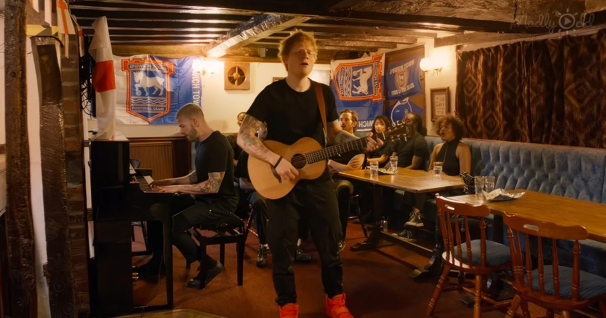 A man plays guitar while another plays piano in a cozy pub setting with patrons seated at tables.