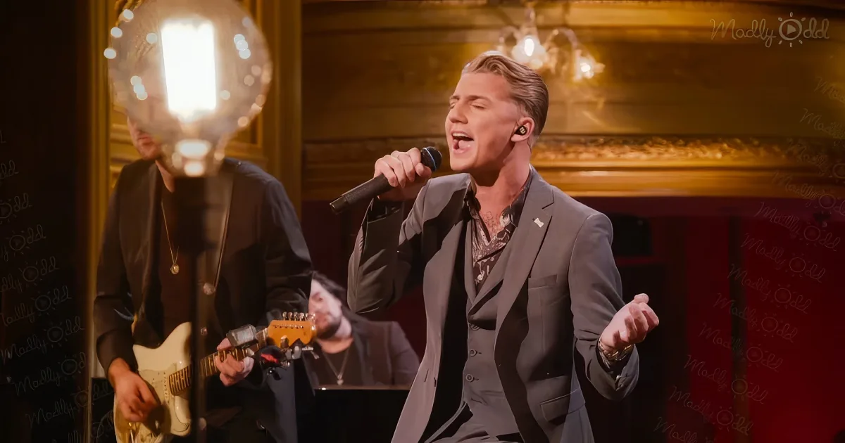 A man in a suit sings into a microphone while playing guitar, with another musician in the background.