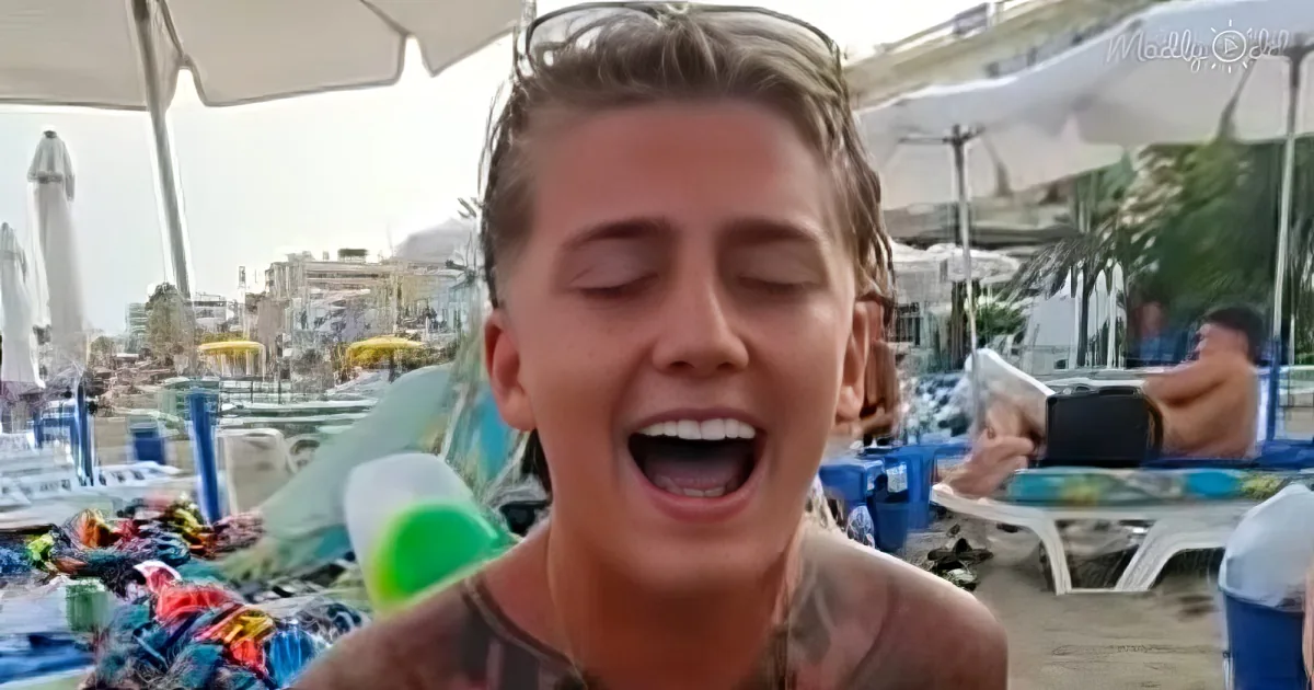 A person with tattoos and sunglasses eats ice cream at a beachside cafe with umbrellas and people in the background.