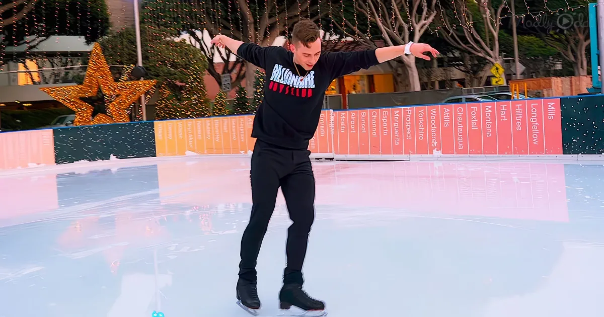 A person in a black outfit skates on an ice rink with colorful barriers and festive decorations in the background.