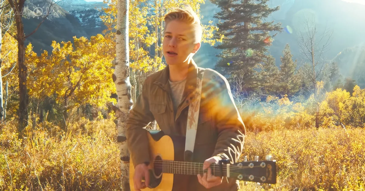 A person plays guitar in a sunlit, autumnal forest with golden leaves and mountains in the background.