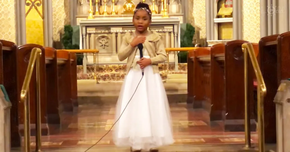 A young girl in a white dress stands in a church, holding a microphone, with her hands clasped near her chest.