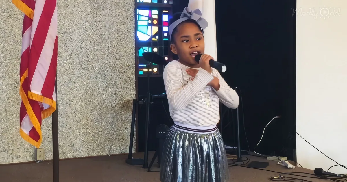 A young girl in a white and silver outfit sings into a microphone on a stage with an American flag and stained glass in...