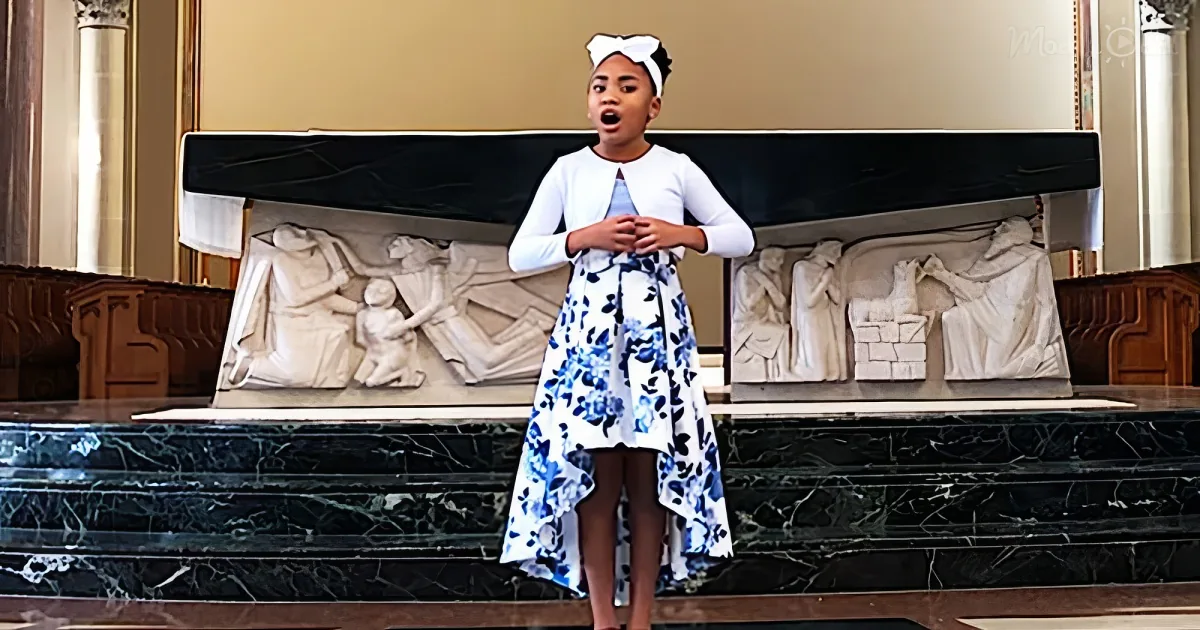 A young girl in a floral dress stands on a marble stage with sculptures behind her.
