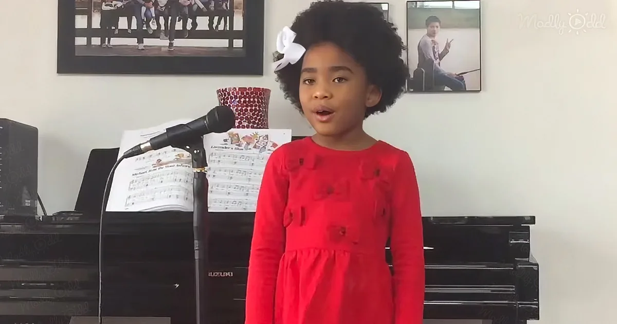 A young girl in a red dress sings into a microphone in front of a piano, with sheet music and framed photos on the wall...