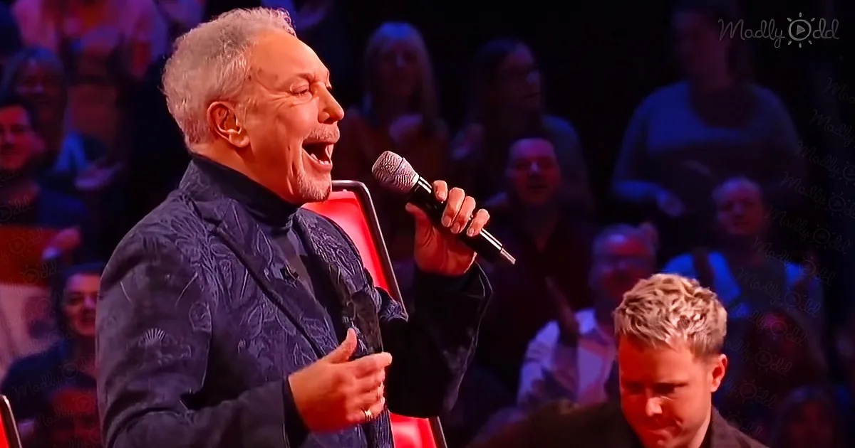 A man in a blue patterned jacket sings into a microphone, gesturing with his right hand.