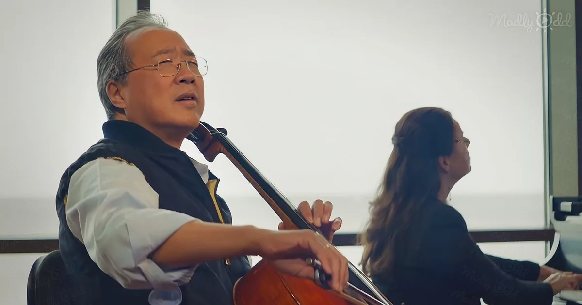An older man in glasses plays a cello, with a woman in the background.
