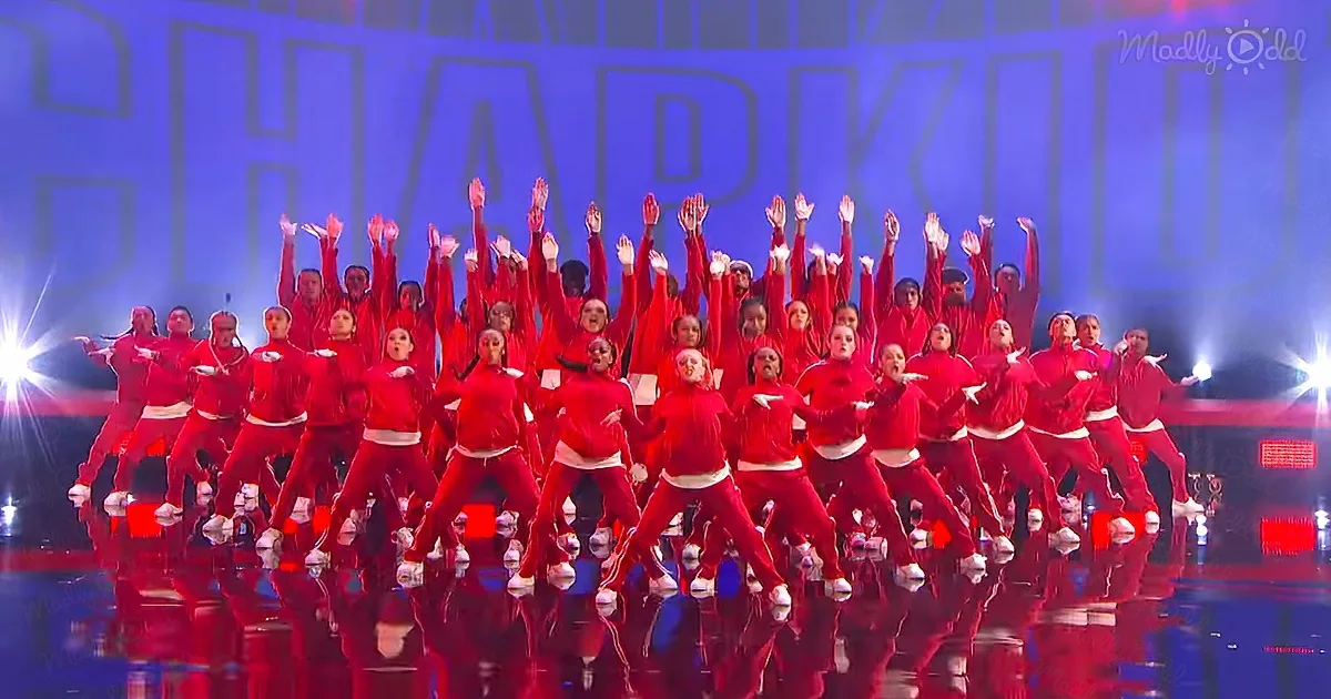 A synchronized dance group in red outfits performs on stage with arms raised.