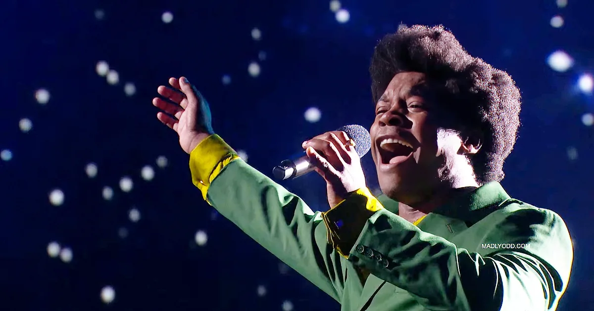 A person in a green outfit sings into a microphone, raising one hand, with a starry background.
