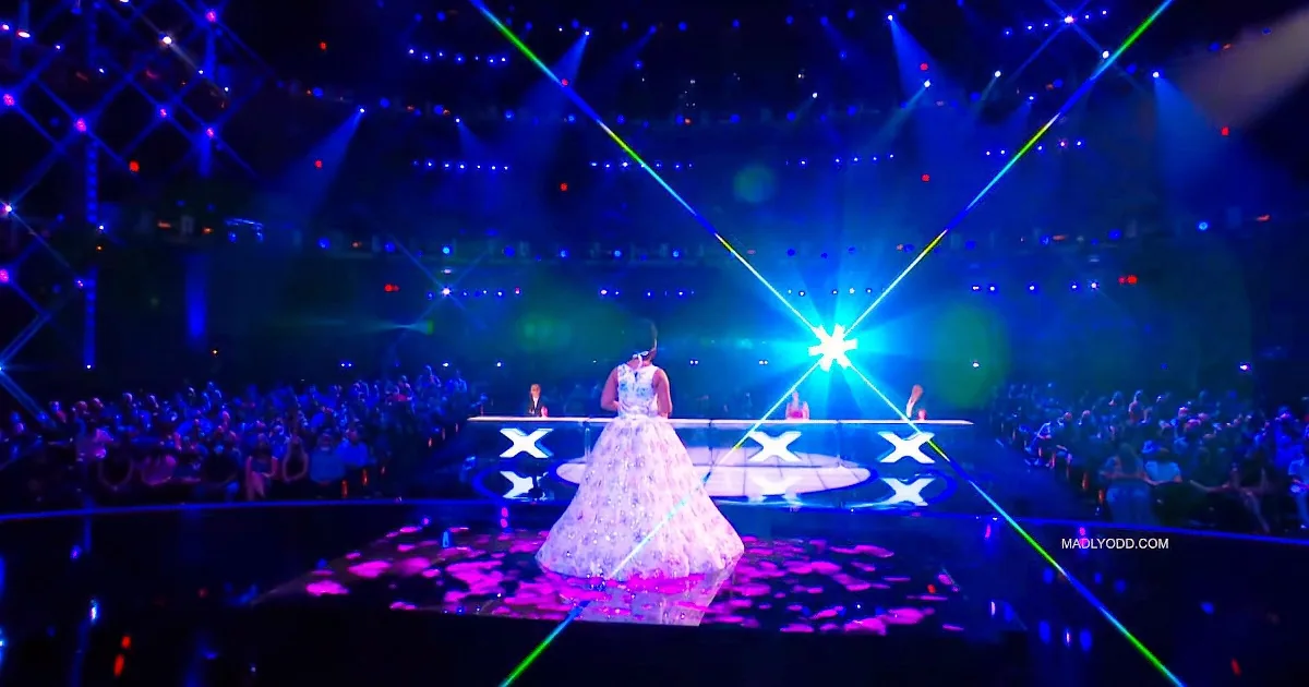 A performer in a sparkling dress stands on a stage with vibrant lights and a large audience.