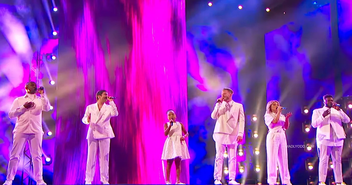 A group of performers in white outfits sing on stage with vibrant purple and pink lighting.