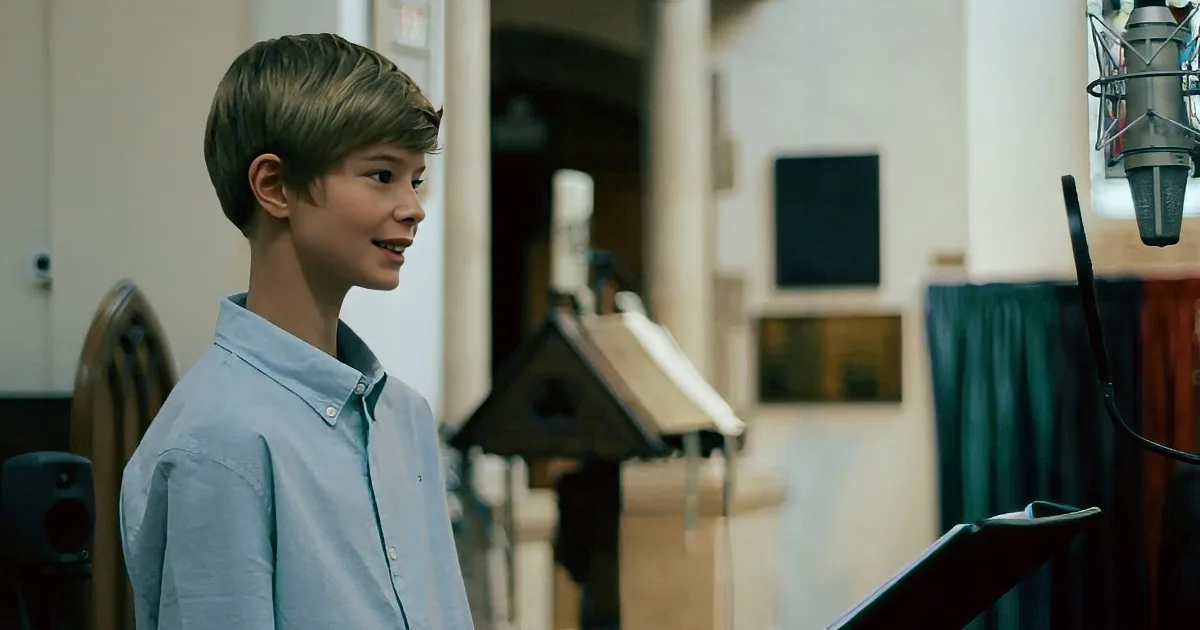 A young boy in a light blue shirt sings passionately in a classroom setting.