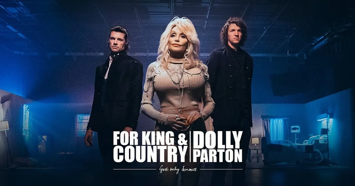 Dolly Parton stands with two men in a room, all holding hands and singing.