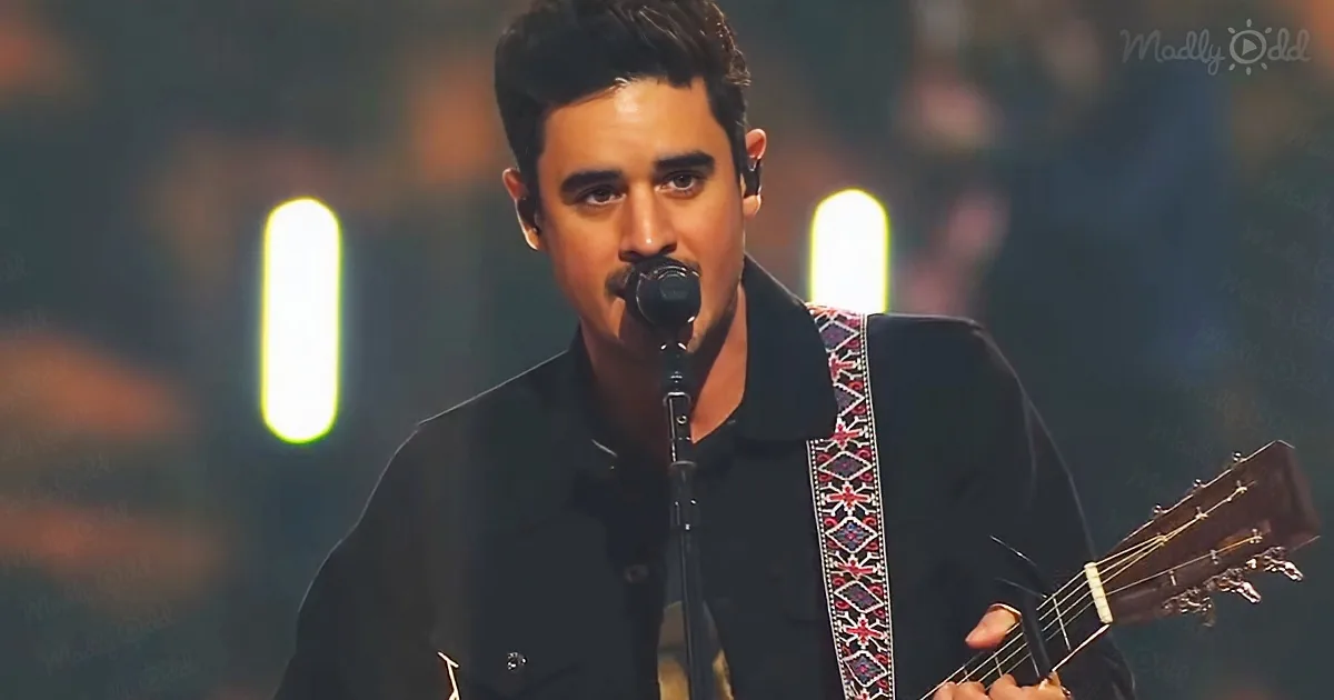 A man in a black jacket plays an acoustic guitar and sings into a microphone on stage.