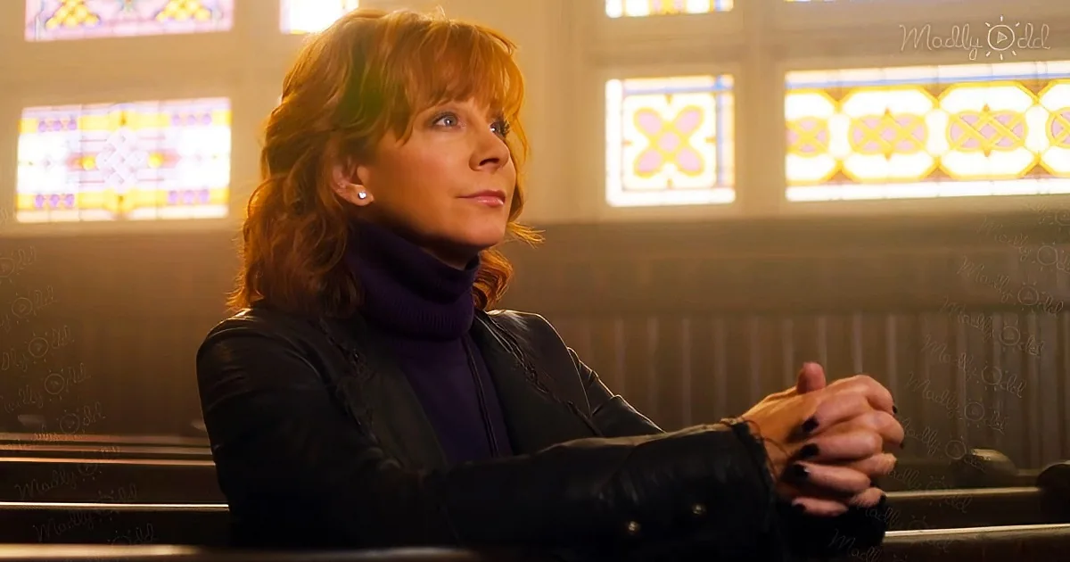 A woman with red hair sits in a church pew, hands clasped, wearing a black jacket and purple turtleneck.