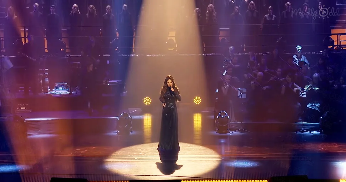 A woman in a black dress stands on a stage with bright lights and an orchestra behind her.
