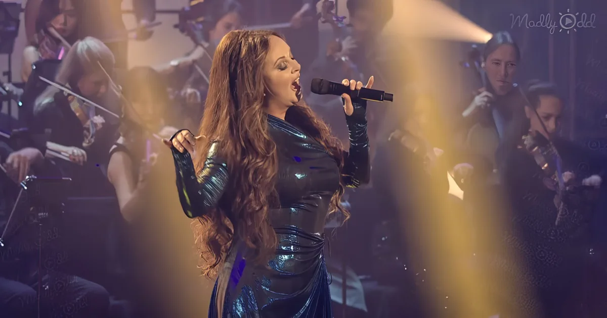 A woman in a shiny outfit sings passionately into a microphone, surrounded by an orchestra.