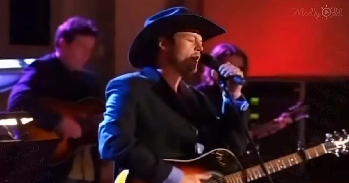 A man in a cowboy hat sings into a microphone while playing guitar, accompanied by other musicians.