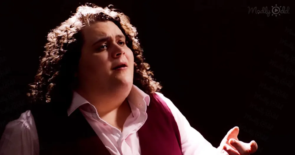 A person with curly hair, wearing a white shirt and red vest, is captured in a close-up shot against a dark background.