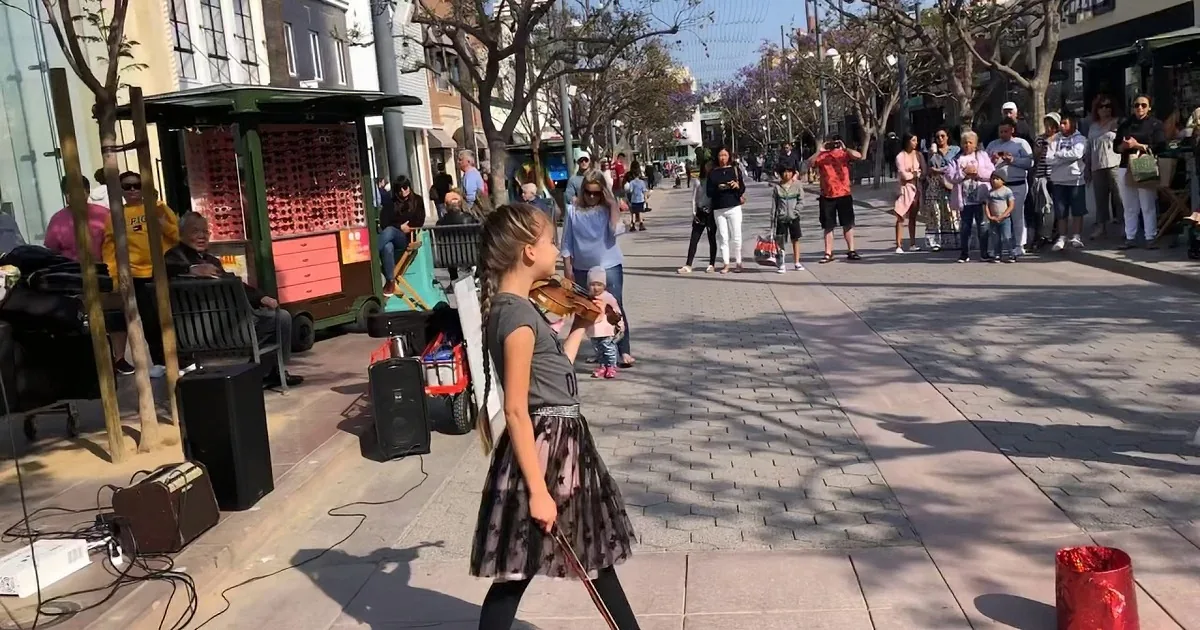 A young girl in a dress plays the violin on a sidewalk, surrounded by street performers and passersby.