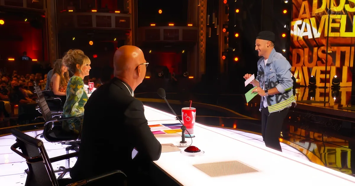 A man in a denim jacket stands on stage, holding papers, while judges sit at a table with red cups.