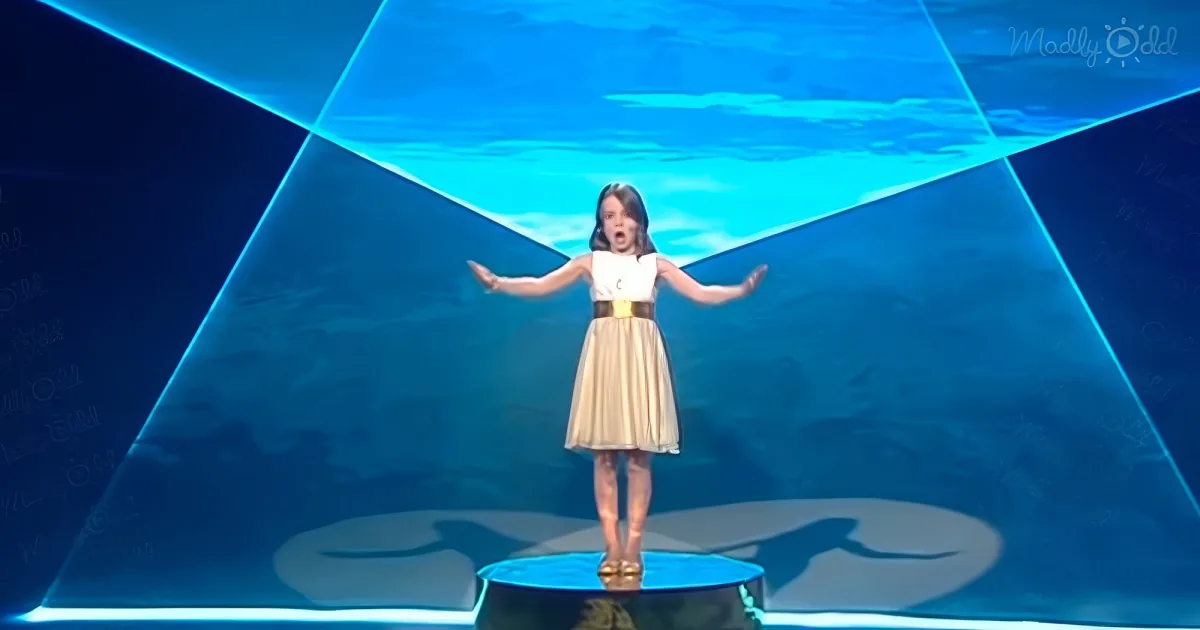 A young girl in a white and gold dress stands on a stage with her arms outstretched, surrounded by a blue geometric...