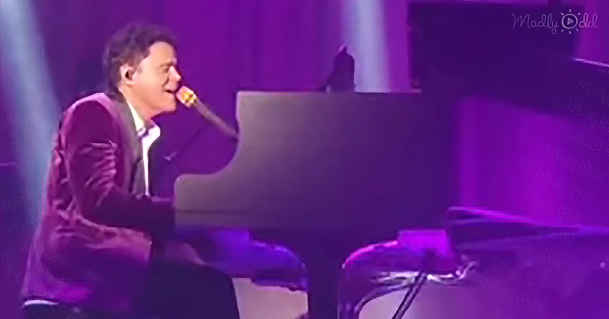 A man in a suit sings into a microphone while playing a piano under purple stage lighting.