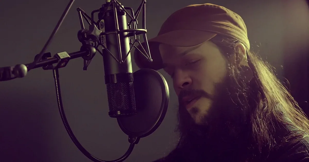 A bearded man in a cap sings into a microphone while holding a guitar, with a "Mesa" amplifier in the background.
