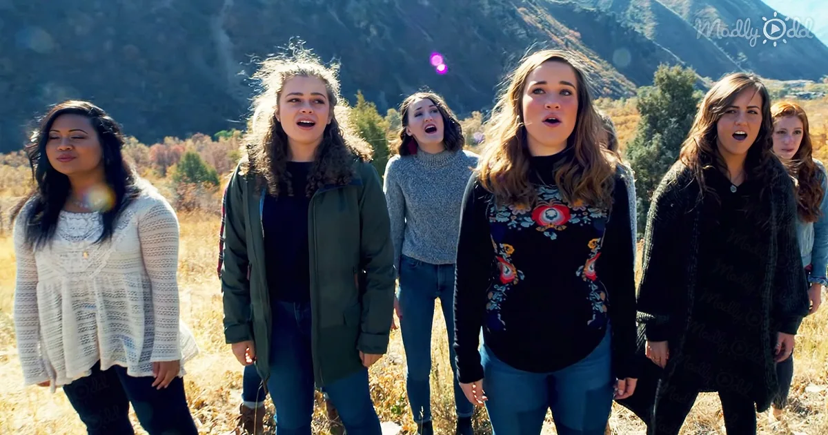 A group of five women sing outdoors in a field with mountains in the background.